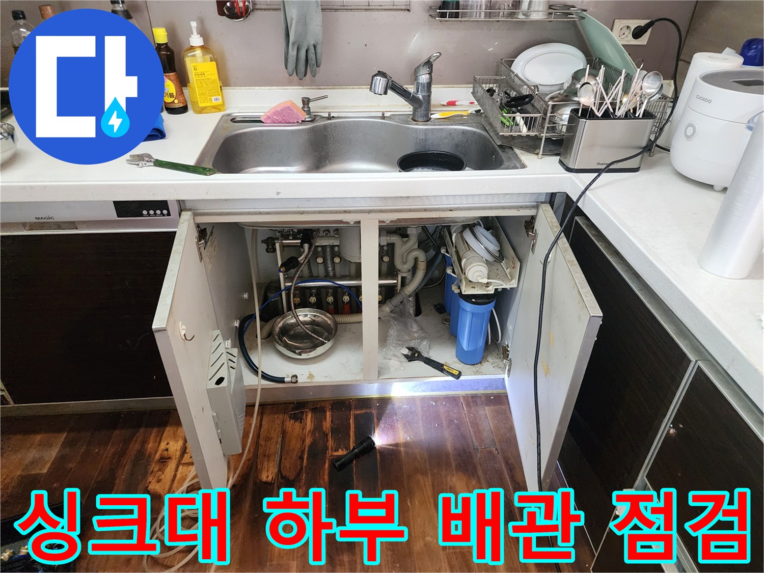 주방 싱크대 하부장 내부의 복잡한 배관과 연결 부위를 손전등으로 비추며 점검하는 모습