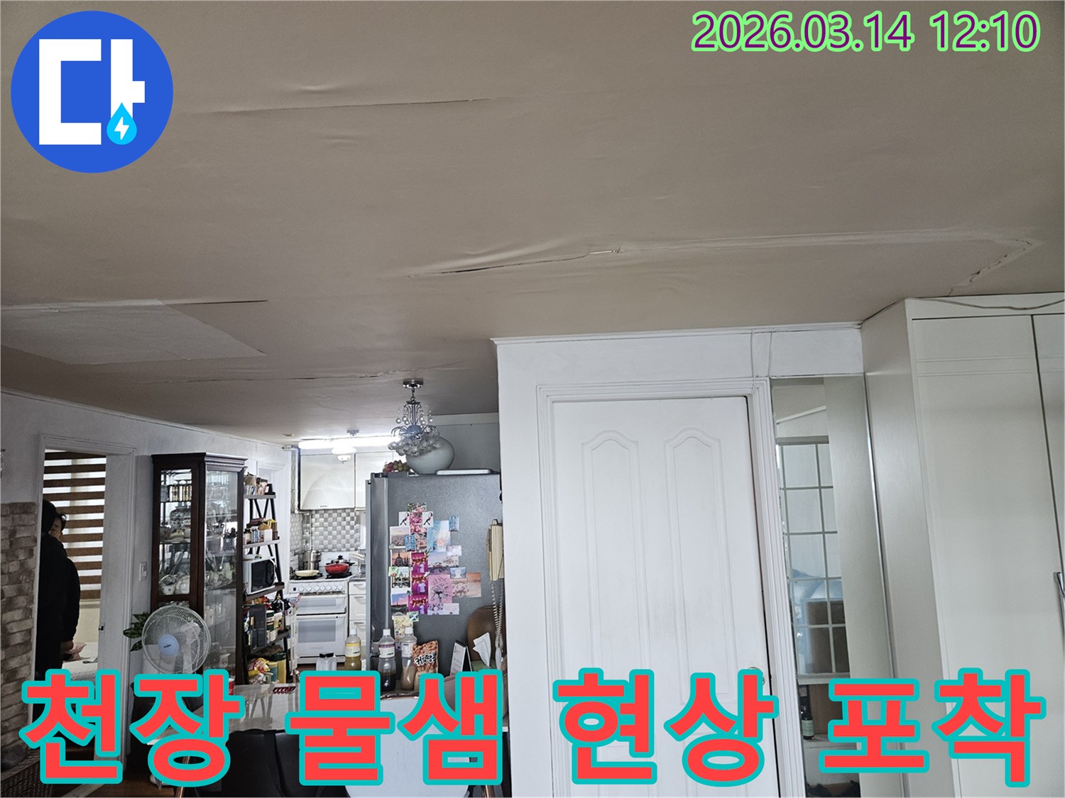 이 사진은 주방과 연결된 거실 천장에 나타난-심각한 물샘 현상을-포착하고 있습니다-천장 도배지가 젖어-늘어지고-변색된 모습은-즉각적인 누수 탐지와-보수 작업이-필요함을-강조합니다