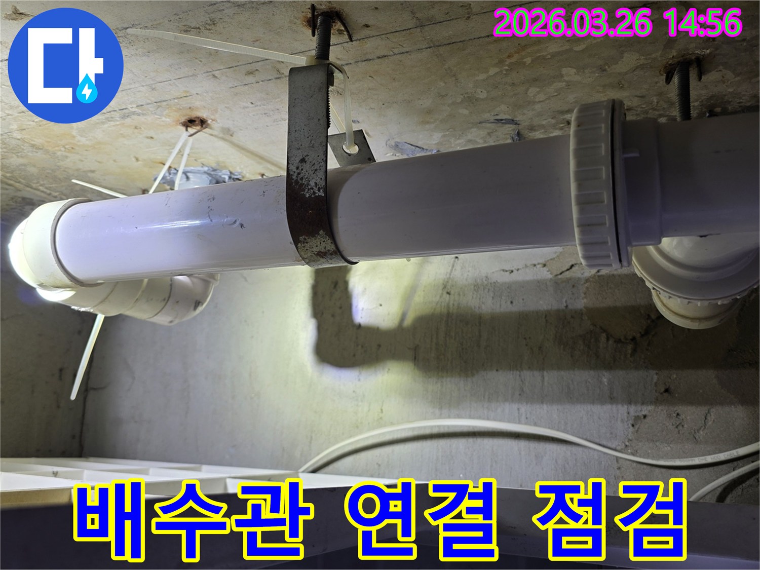 콘크리트 천장 아래에 설치된-하얀색 배수관의 나사식 연결 부위와-이를 고정하는 금속 지지대의 모습
