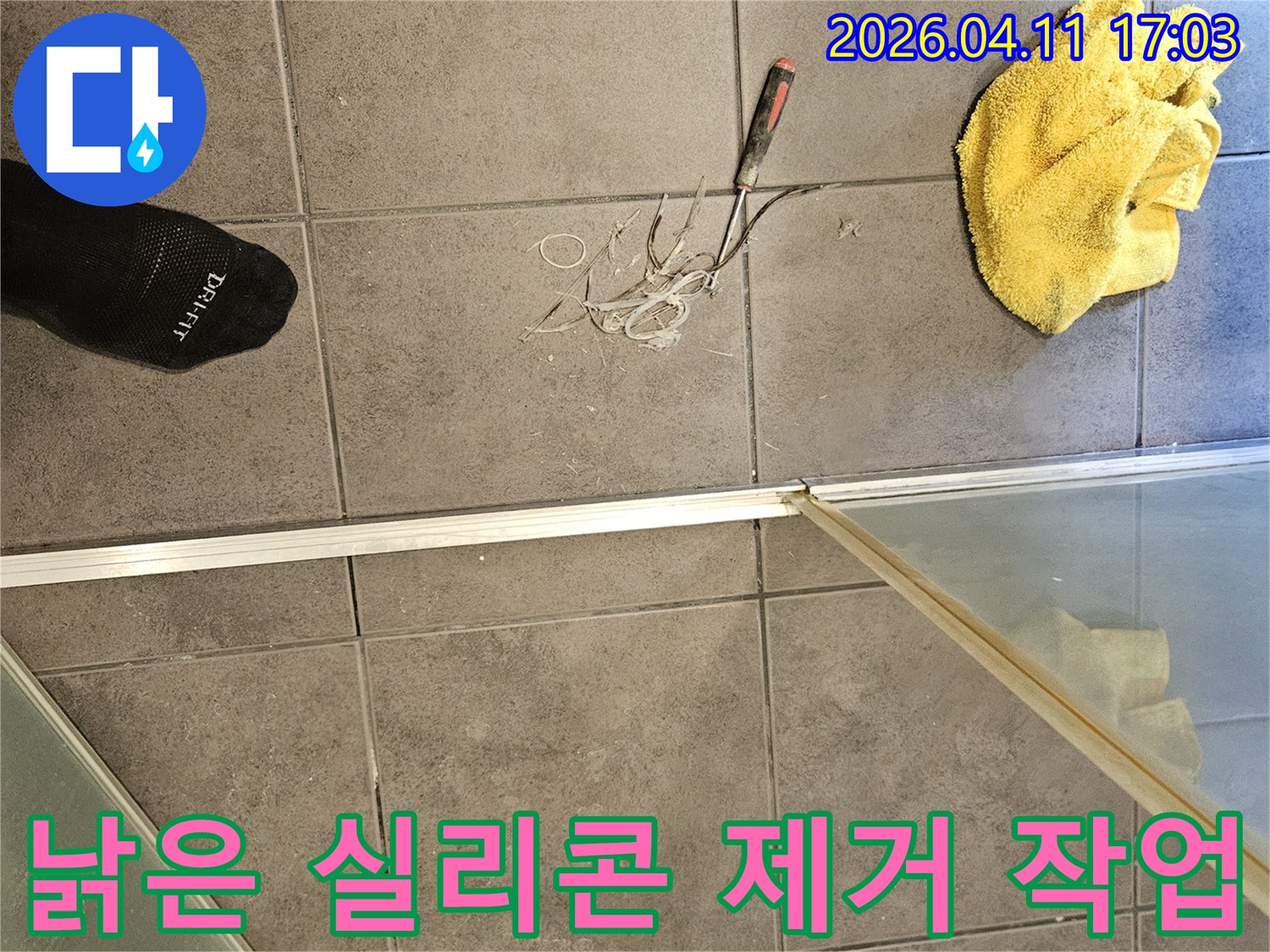 욕실 바닥에 놓인 노란 걸레와-제거된 실리콘 조각들-그리고 샤워부스 경계면 작업 현장