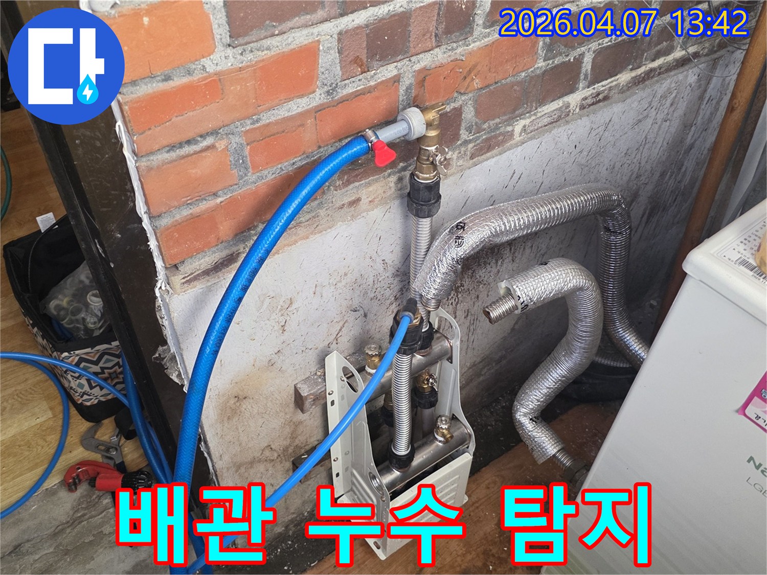 벽돌 벽 옆에-누수 탐지 장비와-파란색 호스가-배관에 연결되어 있으며-누수 지점 확인을 위한-초기 작업이 진행 중입니다.