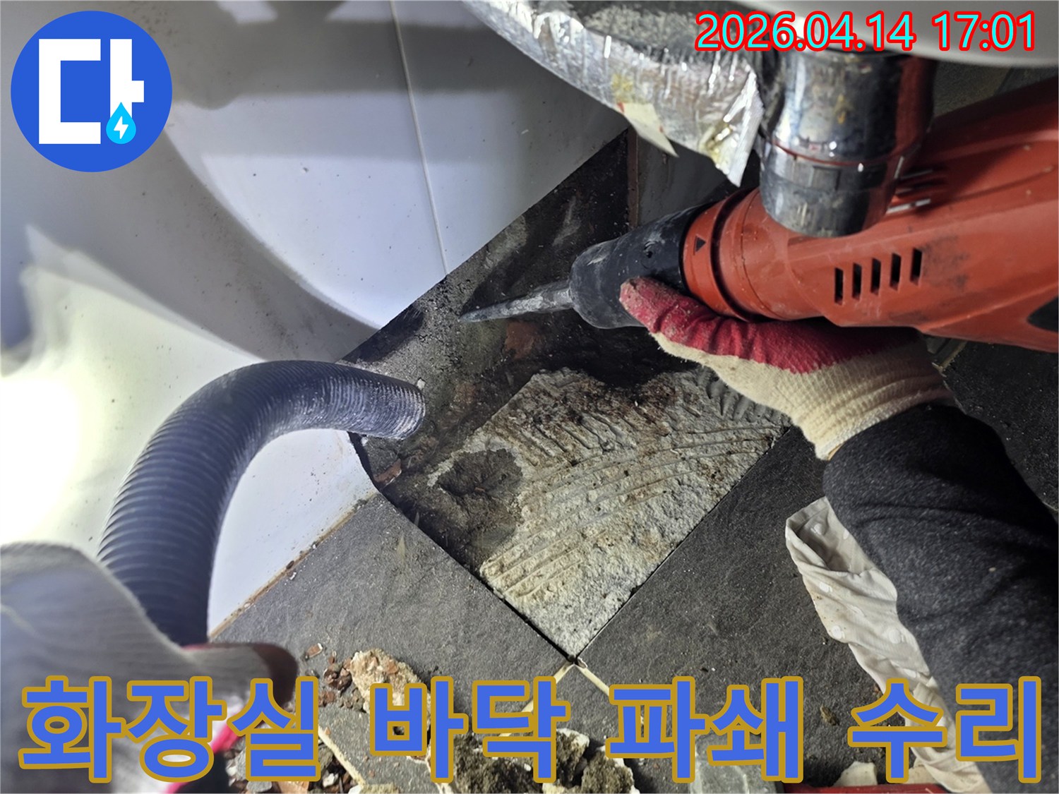 누수 지점 확인 후 화장실 바닥 타일을 파쇄하는 작업자가 드릴을 사용하고-파쇄된 잔해물과 흡입 호스가 보이는 누수 공사 현장