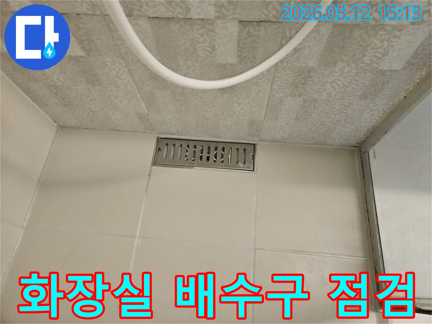 화장실 바닥의 직사각형 스테인리스 배수구와-주변의 흰색 타일-그리고 천장 마감재가 보이는-누수 점검 전 현장 사진입니다.