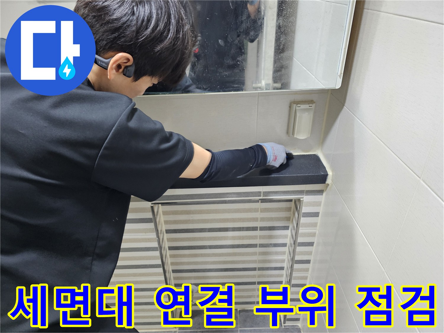 작업자가 세면대 아래쪽의 금속 및 플라스틱 배관 연결 상태를 살피고 있습니다.
