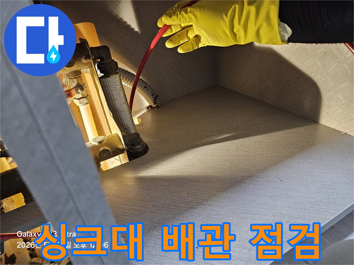 노란 장갑을 낀 작업자가 싱크대 하부의 은색 배관 부위를 확인하고 있습니다.