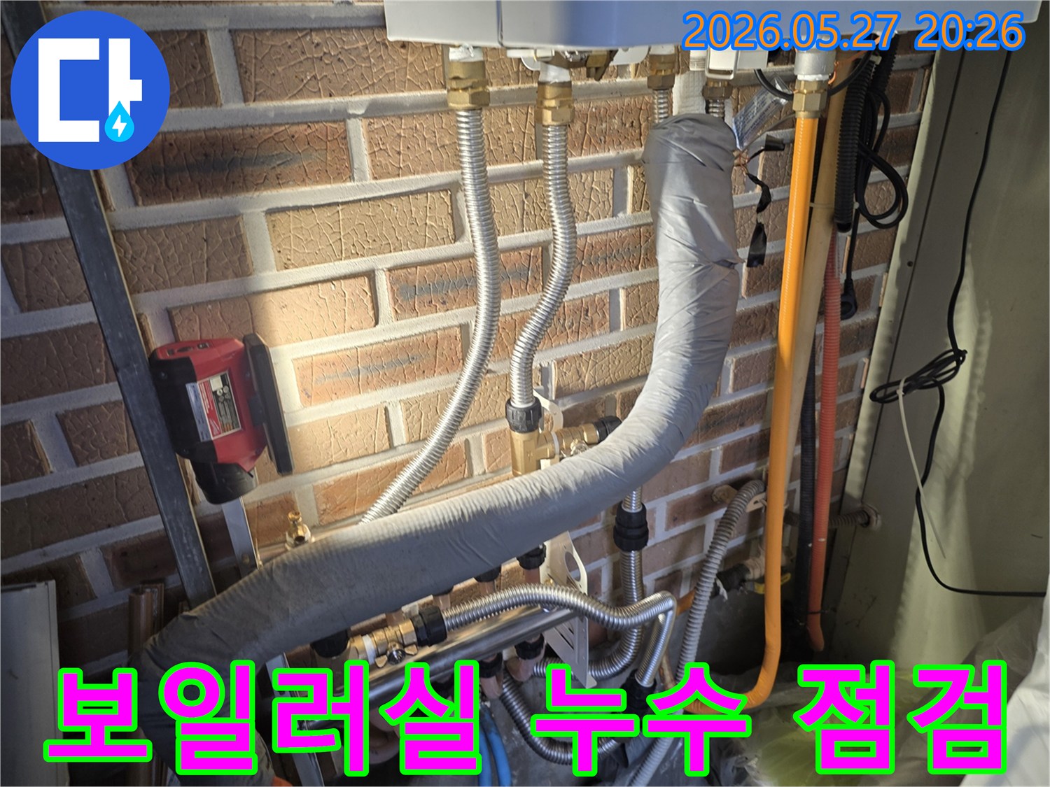 이 이미지는 벽돌 벽에 설치된 보일러 배관과-다양한 연결 부위 그리고 누수 탐지 장비가 보이는 보일러실 현장입니다.