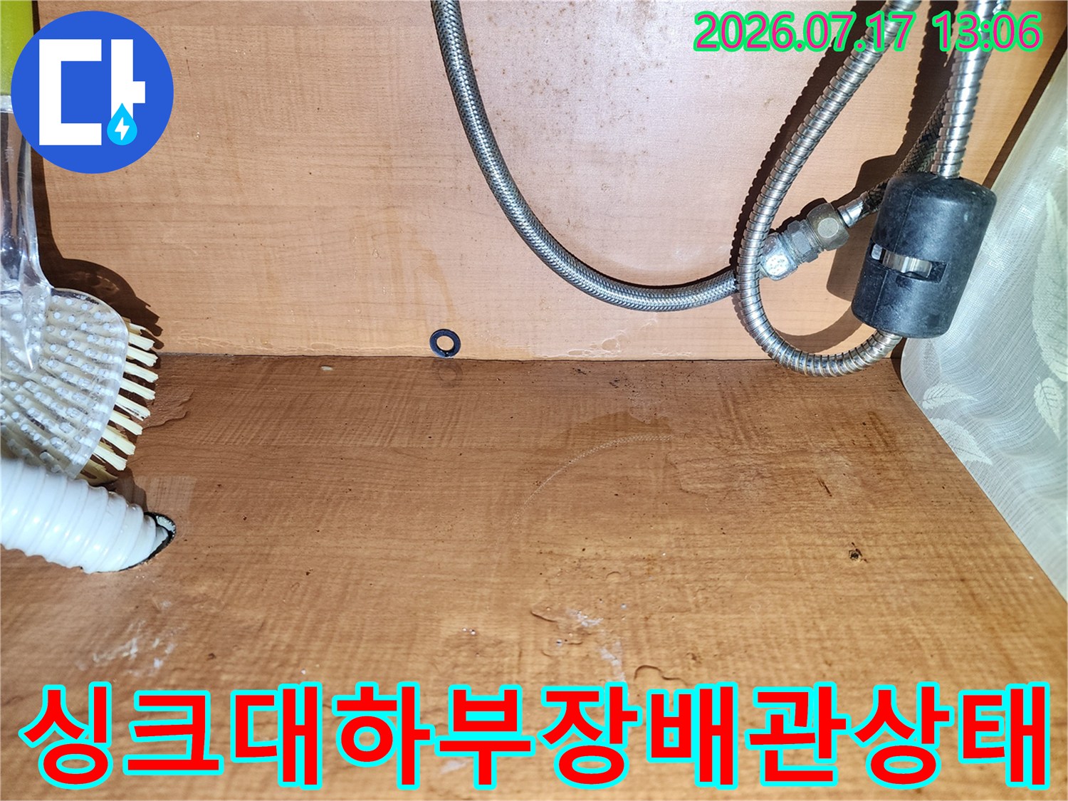 주방 싱크대 하부장의 목재 벽면이 젖어 변색된 모습과 금속제 급수 배관-배수 호스가 설치된 장면
