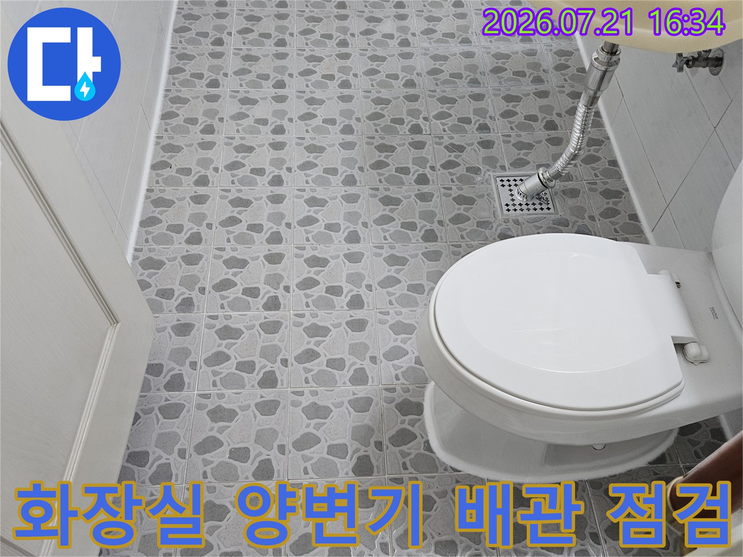 회색 패턴 타일이 시공된 화장실 벽면과 흰색 양변기 아래 노출된 배관 구조물