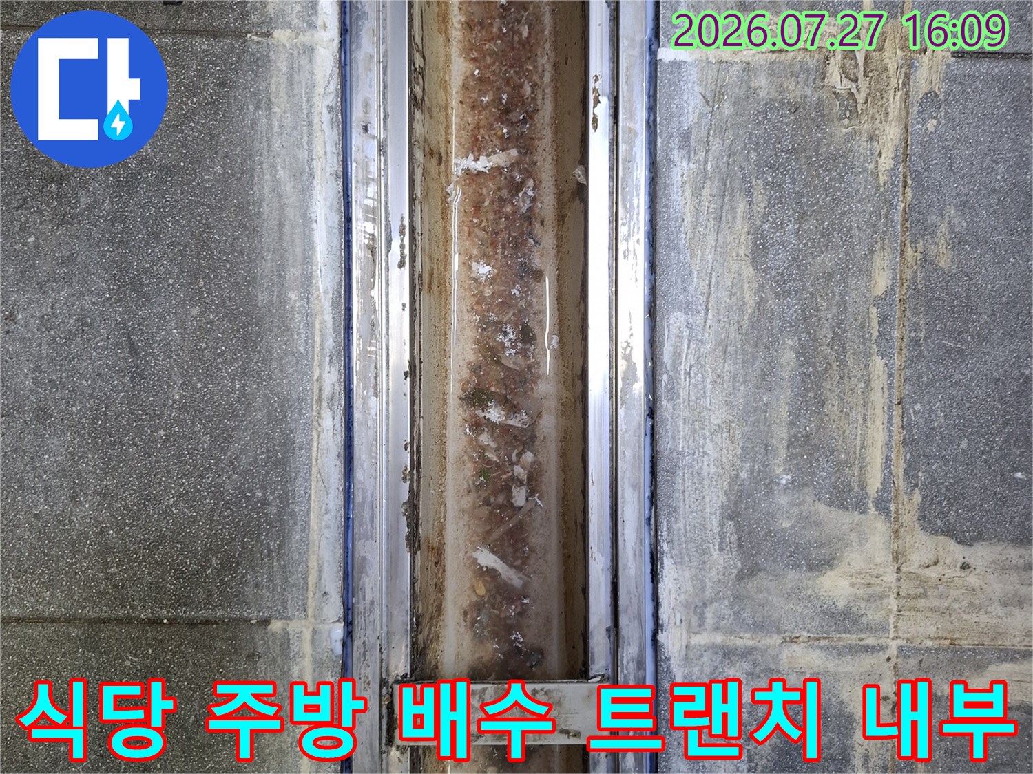 주방 바닥에 설치된 금속제 배수 트랜치의 덮개가 제거되어 내부의 오염물과 방수층 상태가 드러난 모습