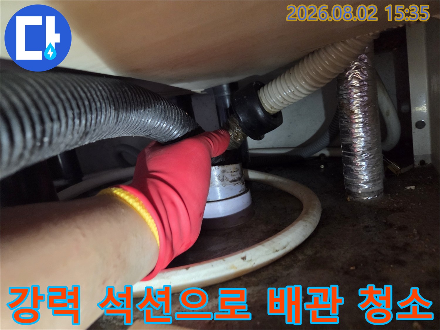 장갑을 낀 손이 싱크대 아래 배수 호스 연결부에 석션기를 대고 청소하는 모습