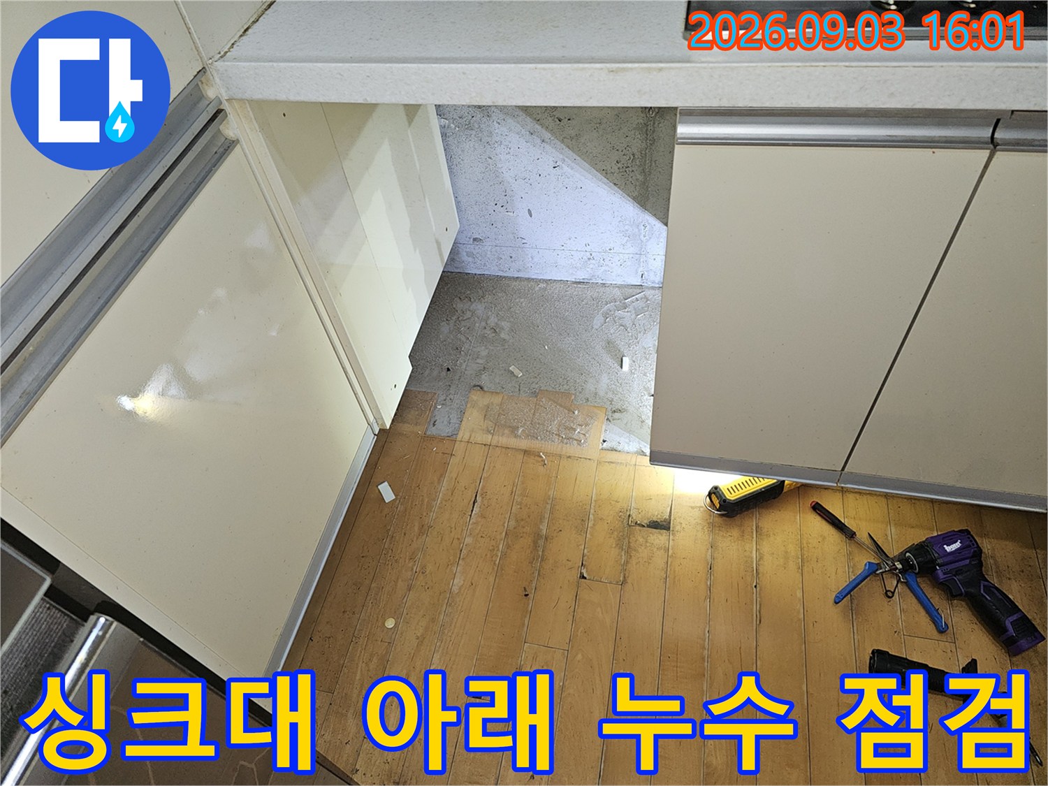 싱크대 하부장 밑 공간에 펜치와 랜턴 등 작업 도구를 배치하고 배관 상태를 확인하는 장면입니다.