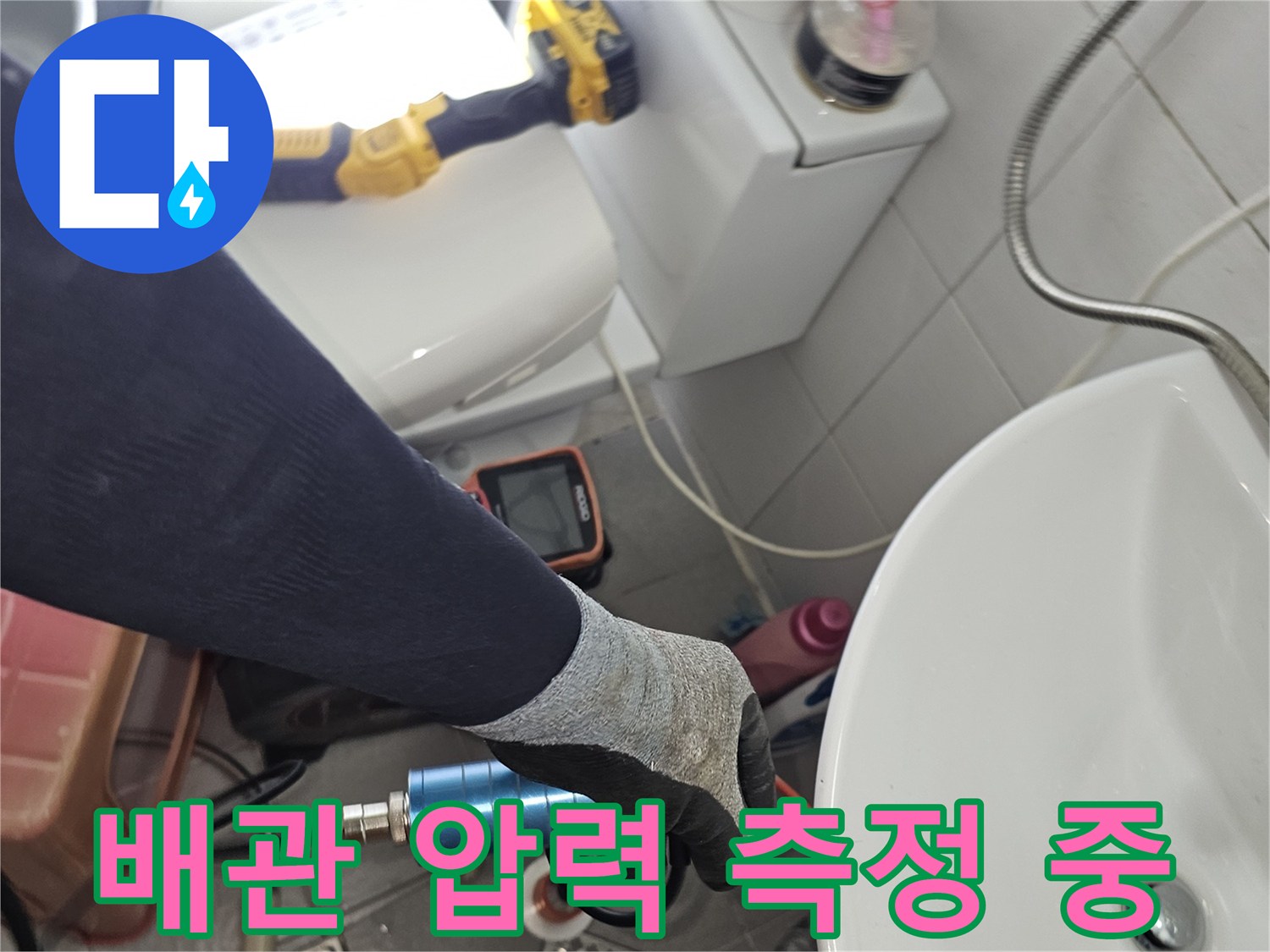 장갑을 낀 작업자가 세면대 아래 배관에 압력계를 연결하는 장면